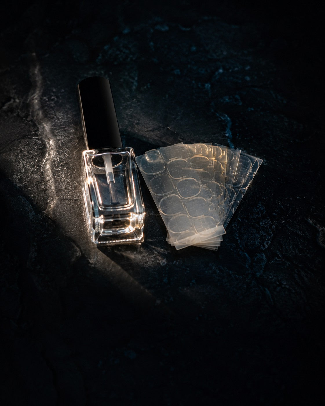 Brush on nail glue bottle beside a fanned stack of adhesive tabs on dark polished stone