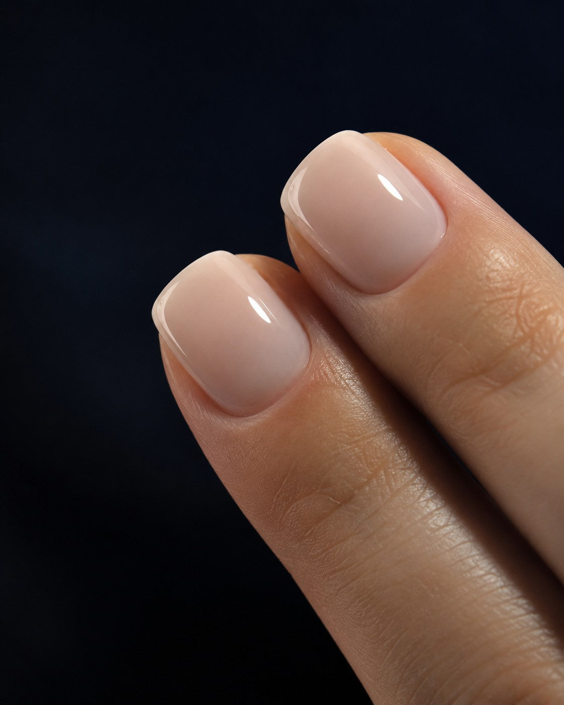 Short squoval press-on nails in a milky nude, shown on two fingertips against a midnight background